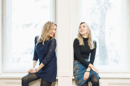 Two blonde female friends chatting and laughing while sitting by the wall between two windows. Breaking down the barrier between people conceptの写真素材