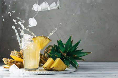 Pineapple fresh juice with falling ice cubes making splash. Cut fruit slices on wooden table and grey concrete background, front viewの写真素材