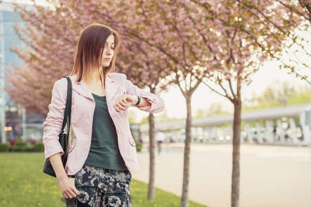 Young woman waiting for the meeting or date and checks time on wrist watches. Beautiful lady standing among blooming sakura trees on the streetの写真素材