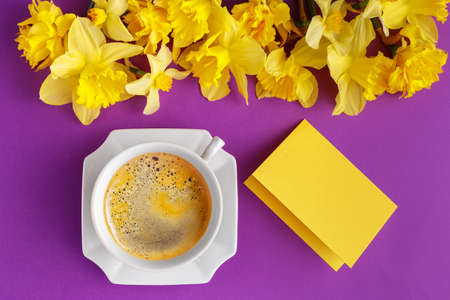 Cup of coffee, empty message card and yellow narcissus flowers on vibrant purple background, spring holiday greetings, top viewの写真素材