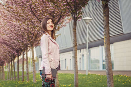 Woman portrait over pink blooming trees in the city street. Beautiful young lady holding her paper notebook near modern buildingの写真素材