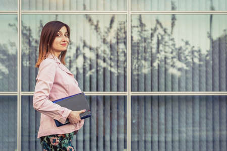 Beautiful young woman portrait holding her paper notebook and laptop near modern buildingの写真素材