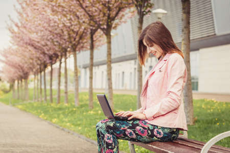 Beautiful young lady sitting on the bench outside and working on laptopの写真素材
