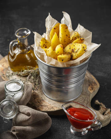 Baked potato wedges in a metal bucket with herbs and tomato sauce on black background - homemade organic vegetable vegetarian potatoes ready to eat snack food mealの写真素材