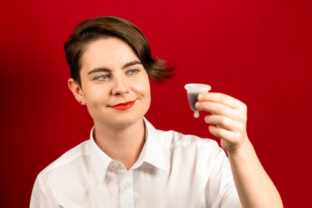 Happy woman holding a menstrual cup - an alternative sanitary product to collect menstrual blood inside, red background, focus on eyesの写真素材
