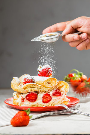 A piece of strawberry cake on a red plate. Man's hand with sieve icing sugar powder from above. Grey background, vertical compositionの写真素材