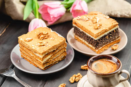 Piece of homemade poppy seed cake and a slice of layered honey cake on the wooden table. Festive set with pastry desserts, cup of coffee and tulip flowers on the backgroundの写真素材