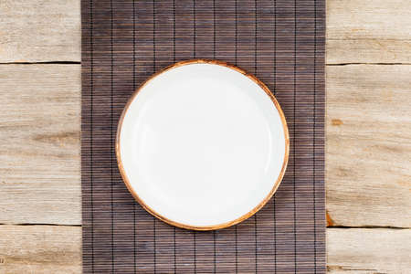 Empty white ceramic dish on rustic wooden table with brown bamboo matの写真素材