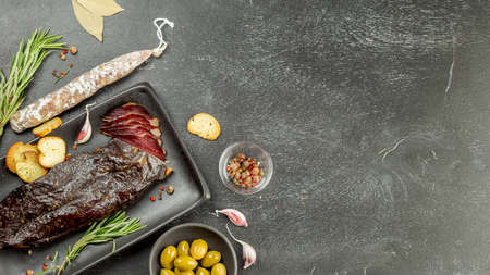 Smoked sliced duck breast and traditional dried sausage on black board with spices, olives, rosemary and bread crackers. Space for textの写真素材