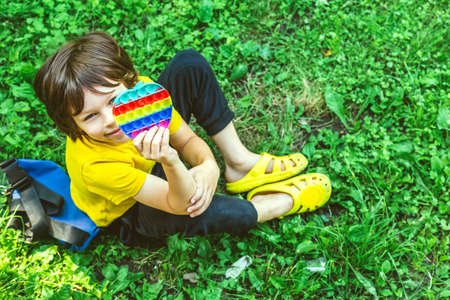 Cute preschooler boy playing with the Pop It Flexible Sensory fidget toy sitting on the grass outdoors. Kids activities, mental development. Selective focus on toy.の写真素材