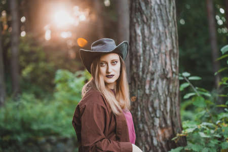 Country style fashion portrait of a blond woman wearing a leather cowboy hat and brown shirt standing in the forest. Dramatic heavy make up with dark lipstick. Halloween costume concept.の写真素材