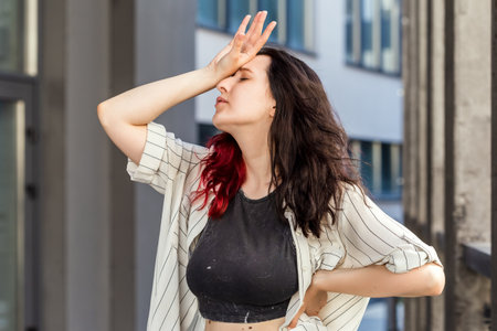 Waist-up shot of white european woman, who is holding arm on forehead showing despair, over office building background. Negative emotion concept. Person who forgot something, poor memoryの写真素材