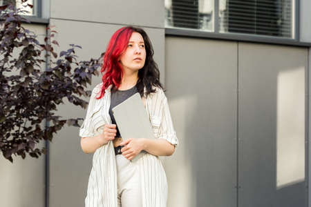 Portrait of a female student or businesswoman with creative red and black hair in smart casual clothing using digital tablet outdoors near the office buildingの写真素材