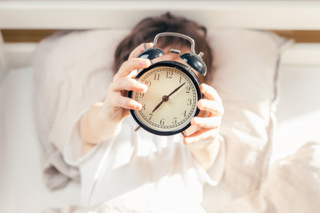 Unrecognizable kid lying in bed and holding a retro style alarm clock. Time to get up in the...