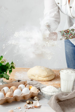 Female Hands Clapping Making Flour Splash above the Dough over Light Background. Eggs and Ingridients are on the Foreground. Empty Space for Textの写真素材