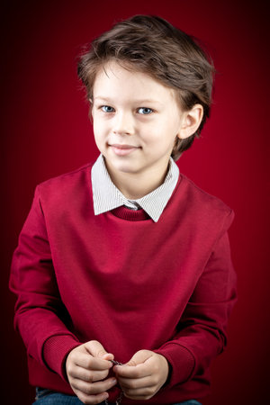 Close up Portrait of Him He Handsome Cute Smiling Smart Caucasian White Boy Dressed in a Red Pullover and Grey Shirt over Dark Red Backgroundの写真素材