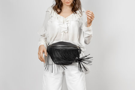 White Woman Wearing White Jeans, Blouse with Frills and Crossbody Black Leather Waist Bag with Decorative Fringe over Grey Backgroundの写真素材