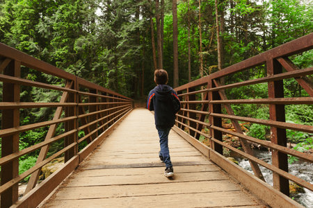 A young boy explores the lush green forest during a summer hike. Kid walking across a rustic wooden bridge, surrounded by tall trees and vibrant foliageの写真素材