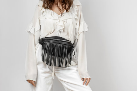 White Woman Wearing White Jeans, Blouse with Frills and Crossbody Black Leather Waist Bag with Decorative Fringe over Grey Backgroundの写真素材