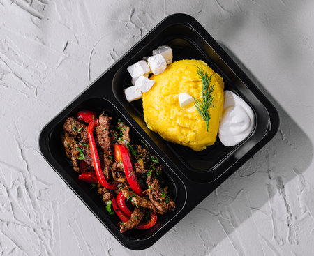 Savory beef stir fry paired with creamy polenta and fresh dill in a black takeout boxの写真素材