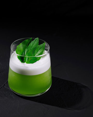 Elegant green cocktail with a frothy top and fresh basil garnish on a dark backdropの写真素材