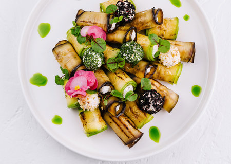 Elegant grilled eggplant slices topped with herbs, flowers, and a drizzle of sauce on a white plateの写真素材