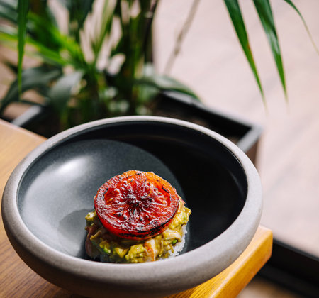 Elegant avocado toast topped with a grilled tomato on a black ceramic plate, presented on a wooden tableの写真素材
