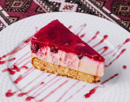 Tempting slice of raspberry cheesecake with syrup on a white plate, over a patterned tableclothの写真素材