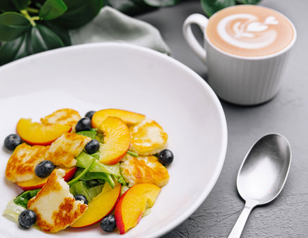 Colorful fruit salad with grilled halloumi, blueberries, and a cup of cappuccino on a dark tableの写真素材