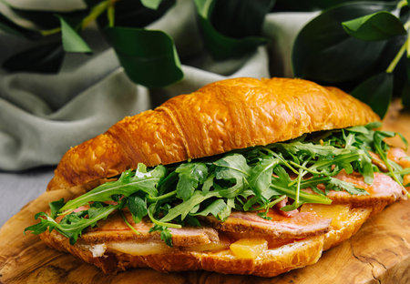 Delicious croissant sandwich filled with bacon, arugula, and cheese on a wooden boardの写真素材