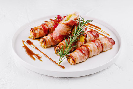Gourmet bacon-wrapped jalapenos filled with cheese, garnished with rosemary on a white plateの写真素材