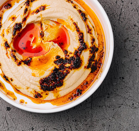 Top view of creamy hummus with chili oil drizzle, served in a white bowl on a textured background