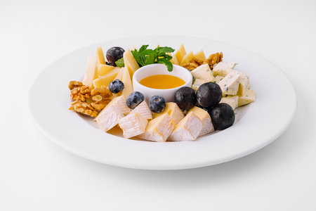 Elegant cheese plate with a variety of cheeses, walnuts, blueberries, and a bowl of honey, isolated on whiteの写真素材