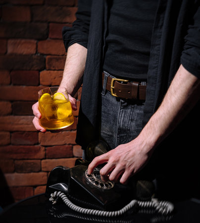 Partial view of a person standing by a table, holding a glass of juice, and dialing an antique phoneの写真素材
