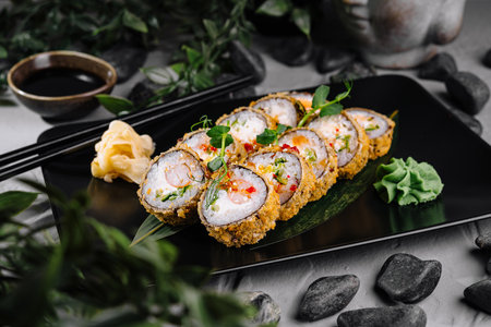 Crispy tempura sushi roll garnished with fresh herbs on a sleek black dish with soy sauce and wasabiの写真素材