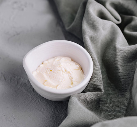 Bowl of creamy mascarpone cheese on a textured grey surface with a green clothの写真素材