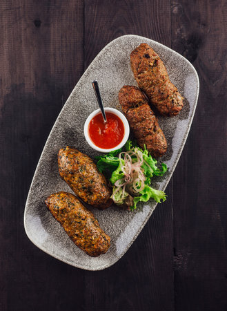 Oval plate with cooked lula kebab seasoned with herbs and spices, decorated with salad and tomato sauce on a dark wooden tableの写真素材