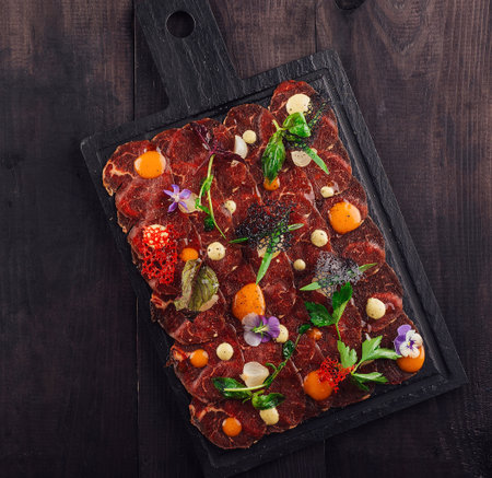 Delicious appetizer of thinly sliced raw beef tenderloin with herbs, flowers and sauce on black board lying on dark wooden tableの写真素材