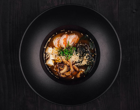Black bowl is filled with a delicious serving of miso soup, complete with tofu, shiitake mushrooms, wakame seaweed, scallions, sesame seeds, and shrimpの写真素材