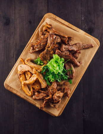 Grilled meat assortment with fresh greens on wooden board, shot from above on dark wooden backgroundの写真素材