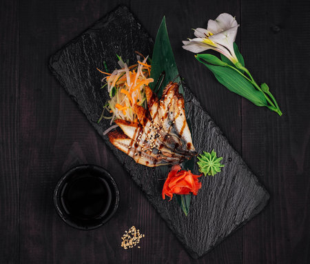 Grilled eel being served on a stone plate with ginger, wasabi, vegetables and a bowl of soy sauce, on a dark wooden tableの写真素材