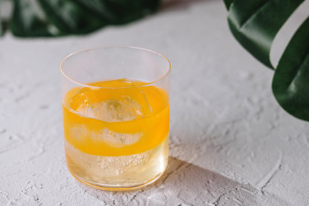 Refreshing yellow cocktail is standing on a white textured background with tropical leavesの写真素材
