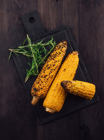 Grilled corn on the cob is lying on a black cutting board and a dark wooden table, decorated with...