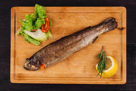 Grilled trout lying on a wooden board with lemon and salad, top view