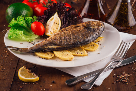Grilled fish served on white plate with potatoes, tomatoes, lemon, lettuce and spices on wooden...