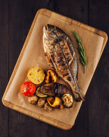 Grilled dorado fish with lemon, rosemary and vegetables on cutting board on dark wooden table