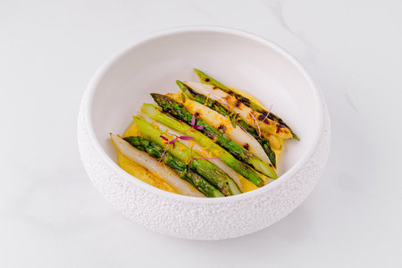 Grilled white and green asparagus arranged on yellow cream with herbs and spices in white bowl on white background, top viewの写真素材