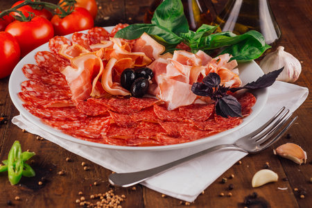 Delicious cold cuts platter with prosciutto, salami, olives and basil leaves is served on a white plate on a wooden tableの写真素材