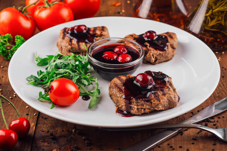 Grilled beef medallions with cherry sauce are served with arugula and cherry tomatoes on a white plateの写真素材