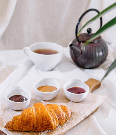 Delicious breakfast with a croissant on a wooden board, accompanied by jam, peanut butter and teaの写真素材
